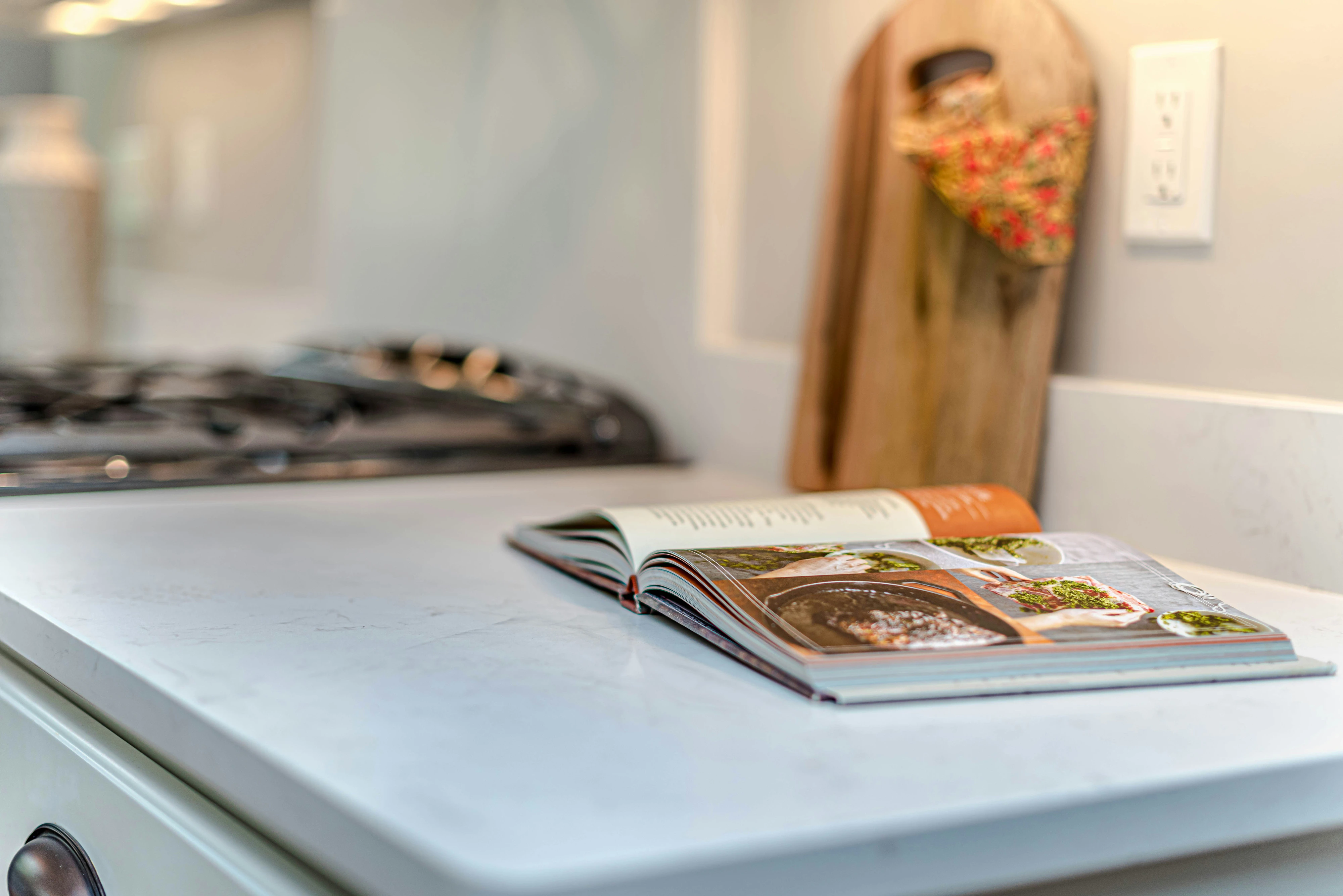 Open cookbook on a white stone countertop beside the range, a wooden board propped against the wall